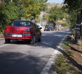 Un vehículo rojo circula por una vía secundaria en el margen derecho una serie de árboles marcados con franjas blancas horizontales