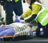 La imagen muestra a un hombre tumbado en una camilla en el suelo con signos de haber sufrido un accidente mientras le atienden los servicios de emergencias