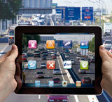 La imagen es un montaje fotográfico en el que dos manos sujetan una tableta donde aparece el icono de varias aplicaciones relacionadas con el tráfico, entre ellas, la de la DGT, y de fondo se ve una carretera con mucha circulación